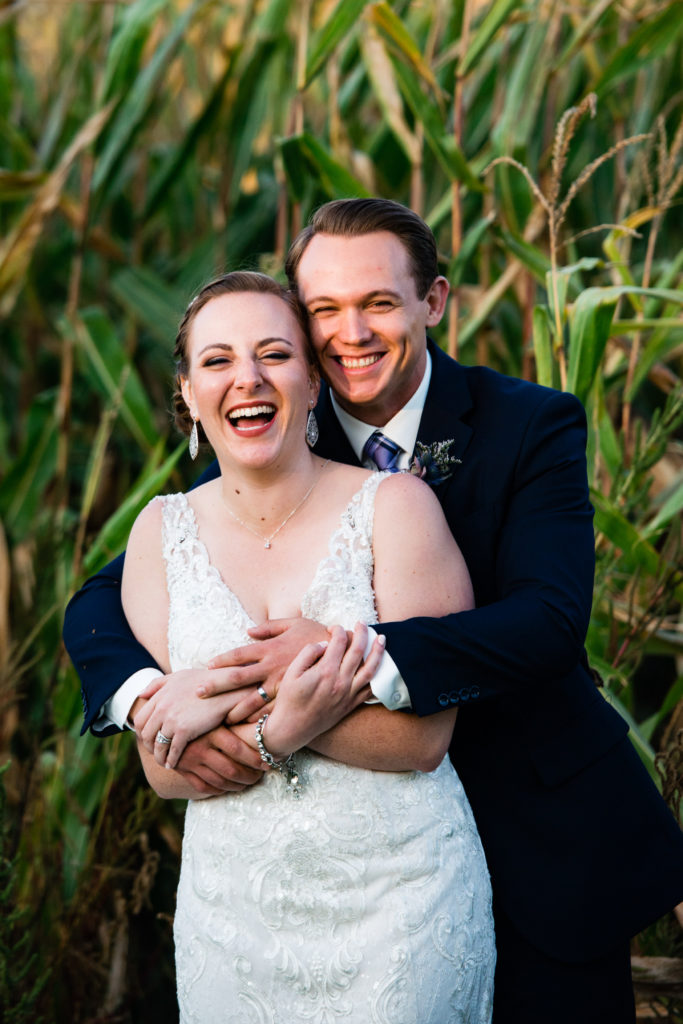 A bride and groom hug and laugh while standing in front of a cornfield.